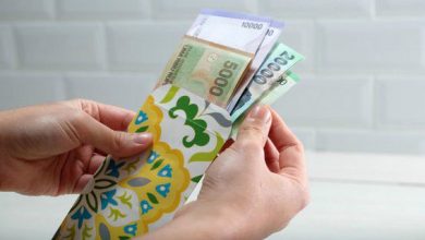 Tangan seseorang sedang memasukkan uang kertas pecahan Rupiah ke dalam amplop THR motif bunga sebagai simbol cara ikhlas bersedekah saat Idulfitri.