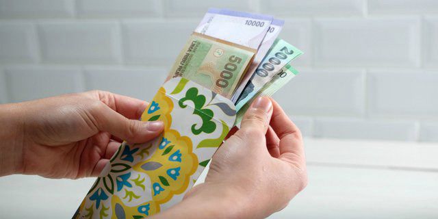 Tangan seseorang sedang memasukkan uang kertas pecahan Rupiah ke dalam amplop THR motif bunga sebagai simbol cara ikhlas bersedekah saat Idulfitri.