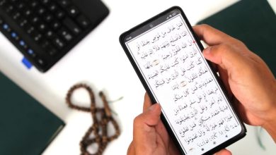 Seseorang memegang ponsel yang menampilkan ayat Al-Qur'an digital dengan latar masjid dan keramaian, menggambarkan digitalisasi tafsir di era modern.