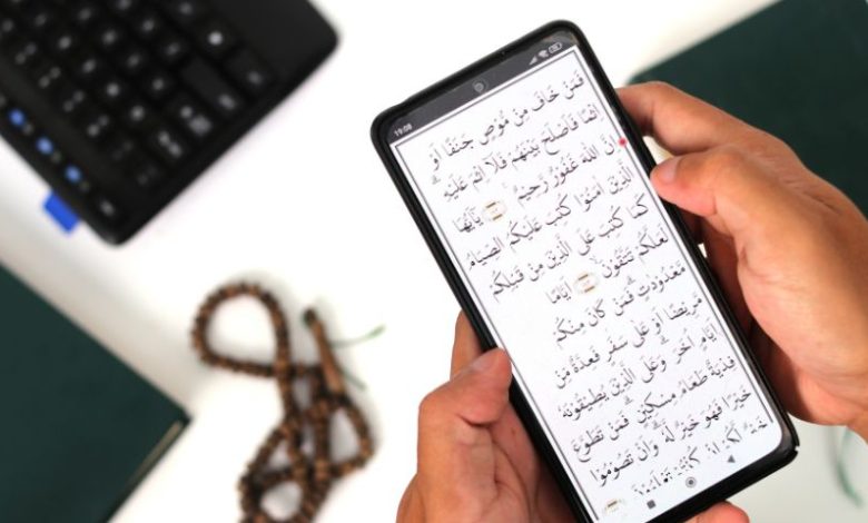Seseorang memegang ponsel yang menampilkan ayat Al-Qur'an digital dengan latar masjid dan keramaian, menggambarkan digitalisasi tafsir di era modern.