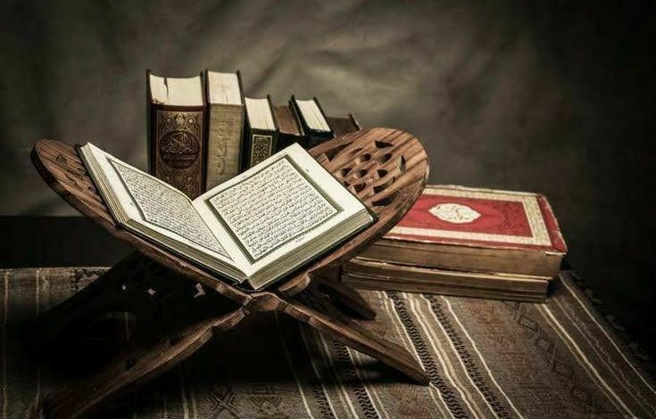 Al-Qur’an terbuka di atas rehal kayu dengan beberapa mushaf di latar belakang
