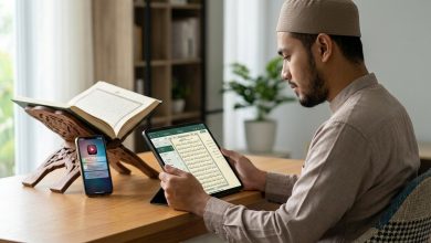 Seorang pria Muslim membaca Al-Qur’an digital melalui tablet di meja, di samping mushaf terbuka dan ponsel dengan notifikasi aktif.