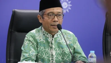 Foto Prof. Hamim Ilyas, Ketua Majelis Tarjih dan Tajdid PP Muhammadiyah, mengenakan baju batik hijau dan peci hitam saat berbicara di depan mikrofon dengan latar belakang logo Muhammadiyah.