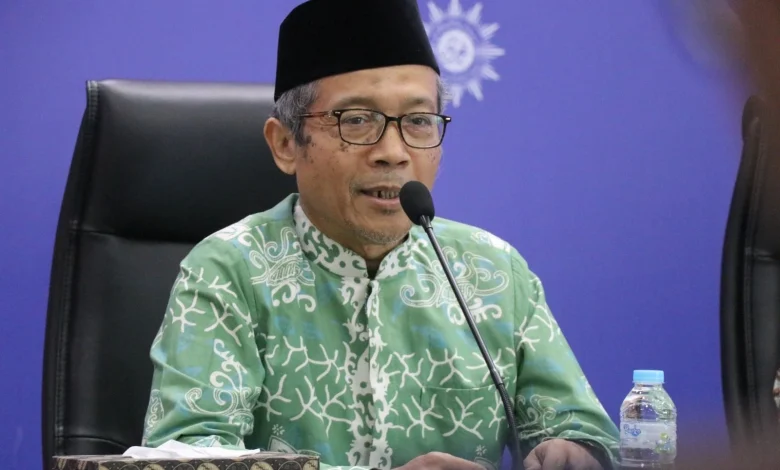Foto Prof. Hamim Ilyas, Ketua Majelis Tarjih dan Tajdid PP Muhammadiyah, mengenakan baju batik hijau dan peci hitam saat berbicara di depan mikrofon dengan latar belakang logo Muhammadiyah.