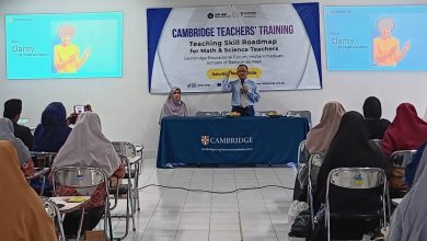 Dua layar presentasi di ruang pelatihan menampilkan slide "Keterjelasan Instruksi" dengan gambar wanita menunjuk. Di antara layar terdapat banner "PELATIHAN GURU CAMBRIDGE: Peta Jalan Keterampilan Mengajar untuk Guru Matematika & Sains". Seorang pembicara pria berbicara dari meja depan sementara penonton, mayoritas berhijab, mendengarkan di ruang kelas yang cerah.