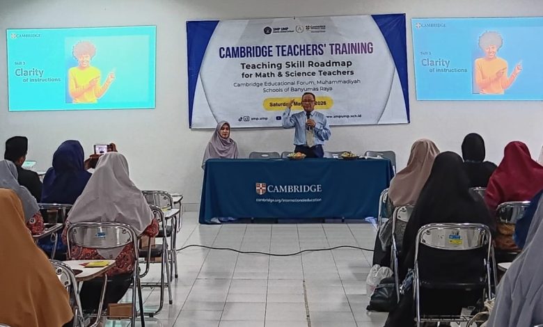 Dua layar presentasi di ruang pelatihan menampilkan slide "Keterjelasan Instruksi" dengan gambar wanita menunjuk. Di antara layar terdapat banner "PELATIHAN GURU CAMBRIDGE: Peta Jalan Keterampilan Mengajar untuk Guru Matematika & Sains". Seorang pembicara pria berbicara dari meja depan sementara penonton, mayoritas berhijab, mendengarkan di ruang kelas yang cerah.