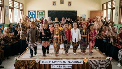 Close-up detail tangan anak TK yang malu-malu memegang ujung baju adat daerah warna-warni, bersiap maju ke panggung sederhana diiringi tepuk tangan guru dan orang tua di latar belakang yang blur.