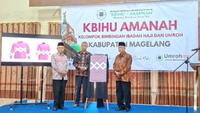 Ketua PDM Kabupaten Magelang dan jajaran KBIHU Amanah menunjukkan contoh seragam batik berwarna pink cerah yang baru diluncurkan dalam sebuah acara seremonial di panggung, lengkap dengan latar spanduk bertuliskan 'KBIHU Amanah Kabupaten Magelang'.