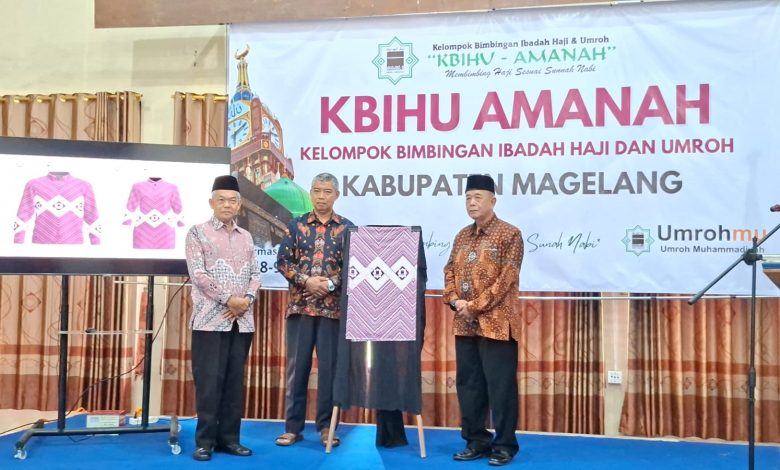Ketua PDM Kabupaten Magelang dan jajaran KBIHU Amanah menunjukkan contoh seragam batik berwarna pink cerah yang baru diluncurkan dalam sebuah acara seremonial di panggung, lengkap dengan latar spanduk bertuliskan 'KBIHU Amanah Kabupaten Magelang'.