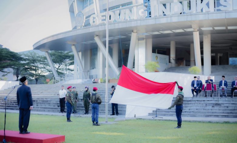 Pimpinan Universitas Muhammadiyah Surakarta (UMS), Prof. Dr. Harun Joko Prayitno, M.Hum., berdiri di mimbar inspektur upacara sambil mengamati petugas pengibar bendera merah putih yang sedang membentangkan bendera besar di halaman Gedung Induk Siti Walidah saat upacara peringatan Hari Pendidikan Nasional 2026. Peserta upacara dan pejabat lainnya duduk di bangku-bangku di latar belakang.