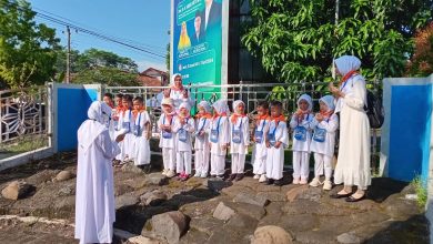 Kelompok siswa TK Aisyiyah se-Kabupaten Pekalongan, mengenakan pakaian ihram dan jilbab putih dengan syal oranye serta tanda pengenal, berdiri di barisan di atas area berbatu. Mereka didampingi oleh tiga guru yang juga mengenakan pakaian putih. Seorang guru di sebelah kiri tampak memberikan instruksi, sementara dua guru di tengah dan kanan berpose dengan tangan terangkat. Mereka semua menghadap ke arah guru di sebelah kiri. Latar belakang menunjukkan pagar besi, tanaman, dan papan reklame yang menampilkan tokoh-tokoh Muslim, di bawah langit biru yang cerah.