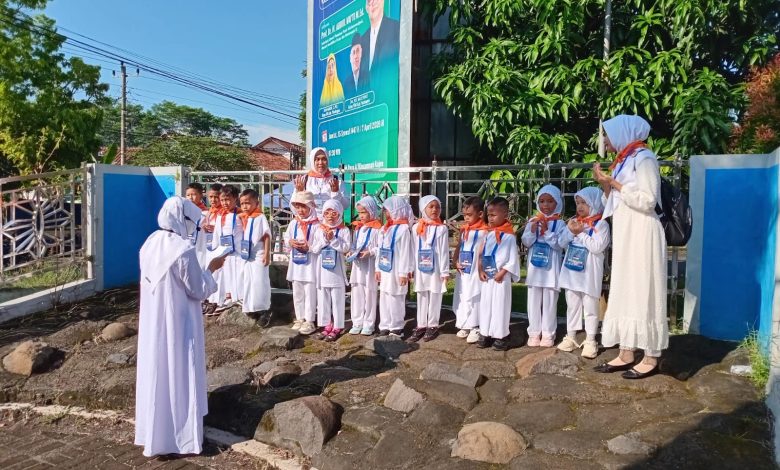 Kelompok siswa TK Aisyiyah se-Kabupaten Pekalongan, mengenakan pakaian ihram dan jilbab putih dengan syal oranye serta tanda pengenal, berdiri di barisan di atas area berbatu. Mereka didampingi oleh tiga guru yang juga mengenakan pakaian putih. Seorang guru di sebelah kiri tampak memberikan instruksi, sementara dua guru di tengah dan kanan berpose dengan tangan terangkat. Mereka semua menghadap ke arah guru di sebelah kiri. Latar belakang menunjukkan pagar besi, tanaman, dan papan reklame yang menampilkan tokoh-tokoh Muslim, di bawah langit biru yang cerah.