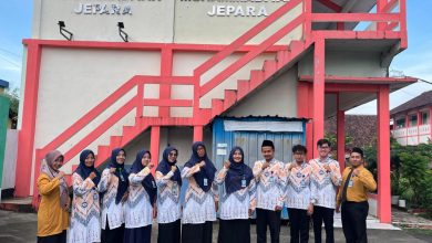 Kelompok guru SMP Muhammadiyah Jepara berpose bersama di panggung terbuka Edutorium K.H. Ahmad Dahlan UMS. Mereka mengenakan seragam batik hijau dan putih berlogo Muhammadiyah serta jilbab hijau. Di tengah, Prof. M. Din Syamsuddin memimpin doa, diapit oleh Rektor UMS Prof. Harun Joko Prayitno dan perwakilan panitia (ketiga dari kanan, memegang piala). Spanduk kecil di latar belakang bertuliskan 'HARDIKNAS SMP MUHAMMADIYAH JEPARA - PENDIDIKAN BERKEMAJUAN & BERAKHLAK MULIA'. Panggung dihiasi tanaman dan bunga. Di latar depan, ratusan siswa SD/MI Muhammadiyah/Aisyiyah, juga mengenakan pakaian ihram dan jilbab putih, duduk khidmat mengikuti doa. Latar belakang menunjukkan tangga Edutorium dan pepohonan hijau di bawah langit biru yang cerah.