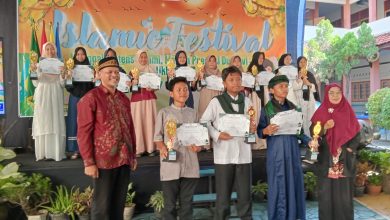 Ibu Amilah, S.Pi. (Kepala SMA Muhammadiyah 1 Pekajangan) berpose bersama dua pemenang utama Islamic Festival Smuhi 2026. Beliau mengenakan jilbab dan kemeja batik hijau Muhammadiyah, tersenyum bangga di tengah, memegang trofi juara umum berbentuk masjid berlapis emas. Di sebelahnya, seorang siswi SMP/MTs (pemenang lomba kaligrafi/qiraat) mengenakan ihram jilbab putih, memegang piala kejuaraan dan sertifikat. Di latar depan, seorang siswa SMP/MTs (pemenang lomba da'i/adzan) mengenakan pakaian ihram putih, berjongkok khidmat, memegang trofi juara dan sertifikat kejuaraan. Mereka berdiri di panggung outdoor Edutorium UMS, di depan spanduk besar 'EDUWISA JEPARA YOGYAKARTA - SMP MUHAMMADIYAH JEPARA', dihiasi tanaman dan bunga. Di latar belakang, ratusan siswa SD/MI Muhammadiyah/Aisyiyah, juga dalam pakaian ihram jilbab putih, duduk khidmat mengikuti doa. Pemandangan Edutorium dan pepohonan hijau di bawah langit biru yang cerah menciptakan suasana hangat, khidmat, penuh kebanggaan, dan sinergi kolektif yang menggembirakan.