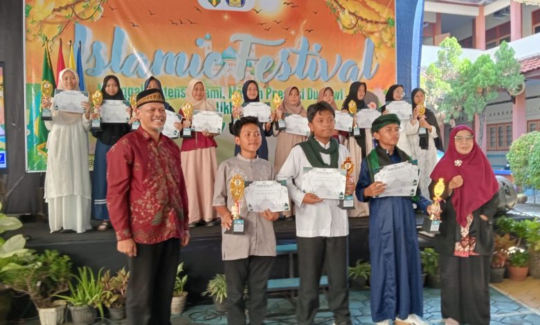 Ibu Amilah, S.Pi. (Kepala SMA Muhammadiyah 1 Pekajangan) berpose bersama dua pemenang utama Islamic Festival Smuhi 2026. Beliau mengenakan jilbab dan kemeja batik hijau Muhammadiyah, tersenyum bangga di tengah, memegang trofi juara umum berbentuk masjid berlapis emas. Di sebelahnya, seorang siswi SMP/MTs (pemenang lomba kaligrafi/qiraat) mengenakan ihram jilbab putih, memegang piala kejuaraan dan sertifikat. Di latar depan, seorang siswa SMP/MTs (pemenang lomba da'i/adzan) mengenakan pakaian ihram putih, berjongkok khidmat, memegang trofi juara dan sertifikat kejuaraan. Mereka berdiri di panggung outdoor Edutorium UMS, di depan spanduk besar 'EDUWISA JEPARA YOGYAKARTA - SMP MUHAMMADIYAH JEPARA', dihiasi tanaman dan bunga. Di latar belakang, ratusan siswa SD/MI Muhammadiyah/Aisyiyah, juga dalam pakaian ihram jilbab putih, duduk khidmat mengikuti doa. Pemandangan Edutorium dan pepohonan hijau di bawah langit biru yang cerah menciptakan suasana hangat, khidmat, penuh kebanggaan, dan sinergi kolektif yang menggembirakan.