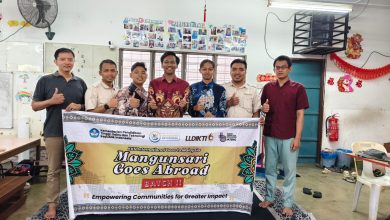 Rombongan mahasiswa KKN Internasional UMS angkatan ke-2 berdiri bangga bersama dosen pendamping dan pengelola Sanggar Bimbingan (SB) Kampung Melayu Sungai Buloh, Malaysia. Ketujuh pria ini tersenyum ke kamera, dengan lima orang di tengah memegang spanduk besar 'KKN INTERNASIONAL GOES TO MALAYSIA - SB KAMPUNG MELAYU SUNGAI BULOH - MANGUNSARI GOES ABROAD'. Mereka mengenakan pakaian rapi, termasuk kemeja batik berlogo matahari Muhammadiyah dan ihram putih jilbab, di bawah cahaya alami Pekalongan. Spanduk besar di latar belakang bertuliskan 'HARDIKNAS SMP MUHAMMADIYAH JEPARA...' dan 'EDUWISA JEPARA YOGYAKARTA...'. Mereka berdiri di panggung outdoor Edutorium UMS, di depan ratusan siswa SD/MI Muhammadiyah/Aisyiyah yang duduk khidmat mengenakan ihram jilbab putih. Pemandangan Edutorium dan pepohonan hijau di latar belakang di bawah langit biru yang cerah menciptakan suasana hangat, bangga, dan penuh kebersamaan yang menggembirakan.