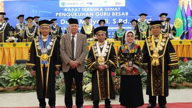 Sesi foto bersama pada acara 'Rapat Terbuka Senat Pengukuhan Guru Besar' di panggung terbuka Edutorium K.H. Ahmad Dahlan UMS. Di tengah panggung, di depan spanduk besar bertuliskan 'SIDANG TERBUKA PROMOSI DOKTOR...', Doktor baru PAI UMS ke-66, Erham Budi Wiranto (memegang toga dan dokumen kelulusan sangat memuaskan, ketiga dari kiri), berdiri diapit oleh promotor Prof. Dr. Waston (mengenakan toga dan seragam batik hijau berlogo matahari Muhammadiyah, kedua dari kiri) dan ko-promotor/sekretaris sidang Dr. Mohamad Ali (mengenakan toga dan batik hijau, keempat dari kiri). Mereka berpose bersama tim narasumber/panitia FIKes UMPP yang memegang piala kejuaraan berbentuk masjid dan sertifikat, serta perwakilan sekolah yang memegang piala. Di latar depan, ratusan siswa SD/MI Muhammadiyah/Aisyiyah duduk khidmat, juga dalam pakaian ihram jilbab putih, mengikuti doa. Latar belakang menunjukkan tangga Edutorium, pepohonan hijau, dan pemandangan modern di bawah langit biru yang cerah. Suasana khidmat, bangga, penuh kebersamaan, dan sinergi kolektif yang menggembirakan.