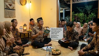 Suasana hangat Pengajian Malam Jumat Karangmangu, Wonosobo, di dalam sebuah rumah warga Javanese yang nyaman. Di tengah, di depan papan tulis portable bertuliskan 'PENGAJIAN MALAM JUMAT KARANGMANGU (SEJAK 1968) - BAITUL ARQOM RANTING - KADERISASI MILITAN - MANHAJ TARJIH', seorang penceramah lokal Muhammadiyah (mengenakan batik dan peci) tersenyum ramah dan memegang buku Manhaj Tarjih, berbicara kepada jemaah. Di latar depan, seorang kader muda (juga batik) duduk khidmat mencatat. Jemaah lain, pria dan wanita (semua berhijab), duduk melingkar mendengarkan dengan serius dan penuh persaudaraan. Ruangan diterangi cahaya hangat lampu gantung dan lampu minyak, menciptakan suasana spiritual, khidmat, dan penuh persaudaraan. Spanduk mading besar di latar belakang bertuliskan 'HARDIKNAS SMP MUHAMMADIYAH JEPARA...' dan 'EDUWISA JEPARA YOGYAKARTA...'.