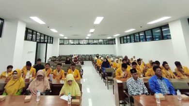 Suasana ruang rapat lantai 3 Gedung FEB UMS yang dipenuhi peserta workshop. Di baris depan duduk tiga ibu pemateri berhijab dan dua bapak pemateri berkemeja. Di belakang mereka, puluhan pelajar IPM SMP MUTU mengenakan jas almamater kuning duduk rapi menyimak acara. Ruangan terang dengan deretan lampu panel dan plafon tinggi dengan unit AC kaset sentral. Terdapat botol air mineral di setiap meja kayu.