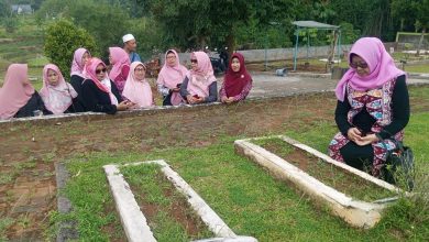 Sebuah foto outdoor yang bersih dan detail dari kader Pimpinan Ranting Aisyiyah (PRA) Karang Tempel dan Pimpinan Cabang Aisyiyah (PCA) Semarang Timur di Pemakaman Muslim Aisyiyah Patemon, Gunungpati. Gambar ini menangkap momen khusyuk ziarah kubur Muhammadiyah, dengan satu wanita berhijab ungu sedang bersimpuh dalam doa di samping dua makam yang sederhana, diapit oleh kelompok kader lain yang berdiri dan memperhatikan dengan penuh hormat. Latar belakangnya adalah bukit, sawah, dan pohon-pohon tropis yang rimbun.