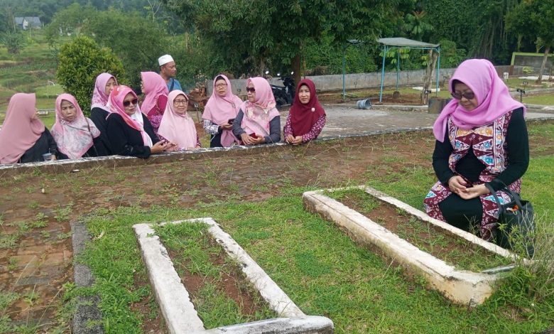 Sebuah foto outdoor yang bersih dan detail dari kader Pimpinan Ranting Aisyiyah (PRA) Karang Tempel dan Pimpinan Cabang Aisyiyah (PCA) Semarang Timur di Pemakaman Muslim Aisyiyah Patemon, Gunungpati. Gambar ini menangkap momen khusyuk ziarah kubur Muhammadiyah, dengan satu wanita berhijab ungu sedang bersimpuh dalam doa di samping dua makam yang sederhana, diapit oleh kelompok kader lain yang berdiri dan memperhatikan dengan penuh hormat. Latar belakangnya adalah bukit, sawah, dan pohon-pohon tropis yang rimbun.