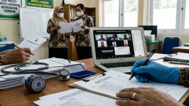 Close-up tangan petugas medis sedang mengisi borang akreditasi klinik pratama dengan latar belakang sesi penilaian daring via laptop dan staf yang berdiskusi di Klinik PKU Muhammadiyah Sapuran.
