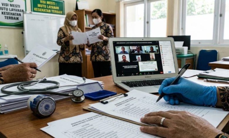 Close-up tangan petugas medis sedang mengisi borang akreditasi klinik pratama dengan latar belakang sesi penilaian daring via laptop dan staf yang berdiskusi di Klinik PKU Muhammadiyah Sapuran.