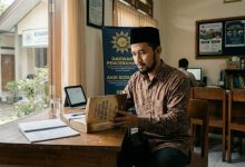 Seorang kader pria Muhammadiyah mengenakan batik dan peci hitam sedang membaca buku hadis di meja kayu kantor komunitas di Purwokerto dengan suasana tenang.