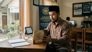 Seorang kader pria Muhammadiyah mengenakan batik dan peci hitam sedang membaca buku hadis di meja kayu kantor komunitas di Purwokerto dengan suasana tenang.