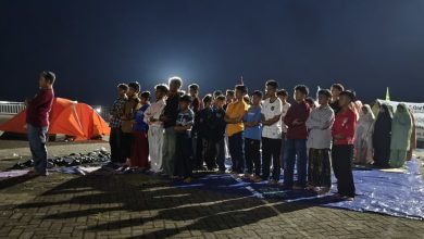 Sekelompok besar siswa pria berbaris di atas tikar biru, dipimpin oleh seorang pemimpin di bagian kiri, bersiap untuk salat di bawah langit malam di lokasi perkemahan perkerasan dengan tenda-tenda oranye.