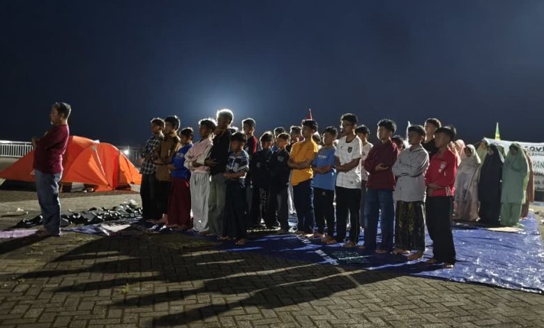 Sekelompok besar siswa pria berbaris di atas tikar biru, dipimpin oleh seorang pemimpin di bagian kiri, bersiap untuk salat di bawah langit malam di lokasi perkemahan perkerasan dengan tenda-tenda oranye.