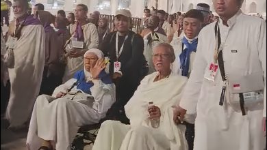 Sebuah foto jurnalisme yang menangkap momen jemaah haji Muhammadiyah di Makkah. Dua jemaah lansia berpakaian ihram putih duduk di kursi roda. Seorang petugas haji Muhammadiyah di sebelah kanan memegang kursi roda. Di latar belakang, ratusan jemaah haji lainnya, termasuk jemaah dengan syal ungu dan biru, memadati area tersebut. Cahaya malam yang hangat menerangi pemandangan tersebut.
