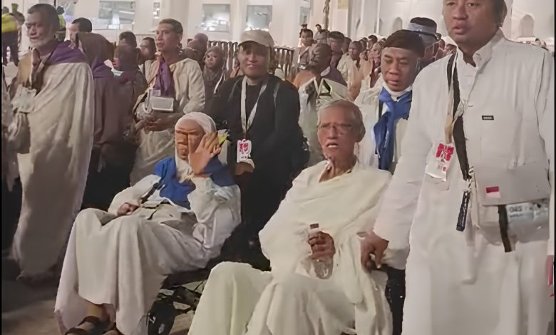 Sebuah foto jurnalisme yang menangkap momen jemaah haji Muhammadiyah di Makkah. Dua jemaah lansia berpakaian ihram putih duduk di kursi roda. Seorang petugas haji Muhammadiyah di sebelah kanan memegang kursi roda. Di latar belakang, ratusan jemaah haji lainnya, termasuk jemaah dengan syal ungu dan biru, memadati area tersebut. Cahaya malam yang hangat menerangi pemandangan tersebut.