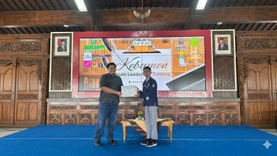 Foto posisi penuh (full shot) memperlihatkan dua orang pria berdiri di atas panggung beralas karpet biru dalam sebuah aula pendopo berarsitektur kayu tradisional. Di sebelah kiri, Wisanggeni Bagus Anggoro memakai kemeja batik hitam lengan pendek dan celana jins biru tua. Di sebelah kanan, seorang pelajar mengenakan seragam OSIS berupa almamater biru tua, celana abu-abu, dan sepatu hitam sedang menyerahkan piagam berbingkai putih. Latar belakang panggung menampilkan layar LED besar bertuliskan 'Kebumen Youth Leadership Training' dengan berbagai logo sponsor di bagian samping.