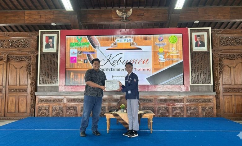 Foto posisi penuh (full shot) memperlihatkan dua orang pria berdiri di atas panggung beralas karpet biru dalam sebuah aula pendopo berarsitektur kayu tradisional. Di sebelah kiri, Wisanggeni Bagus Anggoro memakai kemeja batik hitam lengan pendek dan celana jins biru tua. Di sebelah kanan, seorang pelajar mengenakan seragam OSIS berupa almamater biru tua, celana abu-abu, dan sepatu hitam sedang menyerahkan piagam berbingkai putih. Latar belakang panggung menampilkan layar LED besar bertuliskan 'Kebumen Youth Leadership Training' dengan berbagai logo sponsor di bagian samping.