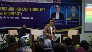 Seorang pembicara pria di atas panggung memberikan presentasi tentang teknologi eDNA untuk konservasi laut dalam sebuah kuliah umum internasional di UNIMUS Semarang. Latar belakang menunjukkan spanduk acara.