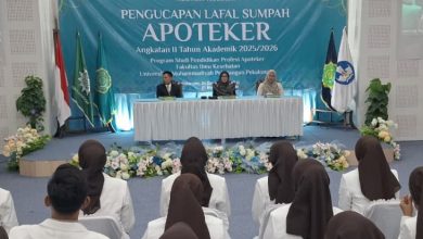 Sekelompok lulusan apoteker UMPP Angkatan II dengan jilbab cokelat dan blazer putih tampak dari belakang, duduk di barisan kursi dengan sarung putih, menghadap ke panggung di mana tiga pejabat duduk di belakang meja yang tertutup kain di depan spanduk besar bertuliskan "PENGUCAPAN LAFAL SUMPAH APOTEKER" dan tiga bendera Indonesia, sedangkan layar proyeksi besar di sebelah kiri dan kanan menampilkan slide dengan foto wajah orang.