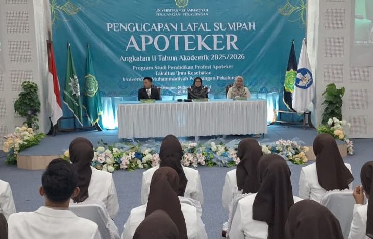 Sekelompok lulusan apoteker UMPP Angkatan II dengan jilbab cokelat dan blazer putih tampak dari belakang, duduk di barisan kursi dengan sarung putih, menghadap ke panggung di mana tiga pejabat duduk di belakang meja yang tertutup kain di depan spanduk besar bertuliskan "PENGUCAPAN LAFAL SUMPAH APOTEKER" dan tiga bendera Indonesia, sedangkan layar proyeksi besar di sebelah kiri dan kanan menampilkan slide dengan foto wajah orang.