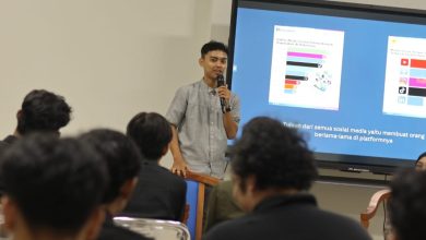 Seorang pemuda pembicara talkshow, Yusuf Susanto, sedang berdiri memegang mikrofon di depan layar presentasi besar. Layar tersebut menampilkan infografis logo media sosial seperti YouTube, Instagram, dan TikTok, serta teks kesimpulan: "Tujuan dari semua sosial media yaitu membuat orang berlama-lama di platformnya." Di depannya duduk beberapa peserta seminar dengan latar belakang kursi perak.
