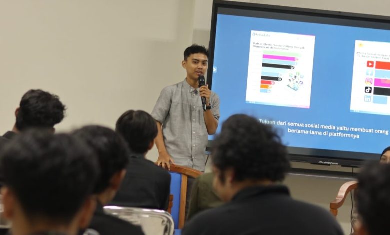 Seorang pemuda pembicara talkshow, Yusuf Susanto, sedang berdiri memegang mikrofon di depan layar presentasi besar. Layar tersebut menampilkan infografis logo media sosial seperti YouTube, Instagram, dan TikTok, serta teks kesimpulan: "Tujuan dari semua sosial media yaitu membuat orang berlama-lama di platformnya." Di depannya duduk beberapa peserta seminar dengan latar belakang kursi perak.