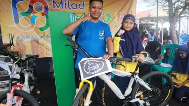 Seorang pria dan anak perempuan berhijab berpose di panggung acara Milad ke-69, memegang hadiah sepeda baru berwarna kuning dan hitam yang masih dibungkus.