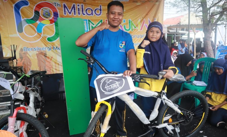 Seorang pria dan anak perempuan berhijab berpose di panggung acara Milad ke-69, memegang hadiah sepeda baru berwarna kuning dan hitam yang masih dibungkus.
