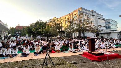 Sejumlah pria, termasuk remaja dan anak-anak, duduk rapi di atas sajadah di area parkir paving yang luas untuk sholat, dengan mimbar kayu kecil di karpet merah di depannya, di luar ruangan dekat gedung-gedung modern dan pohon di pagi hari yang cerah.