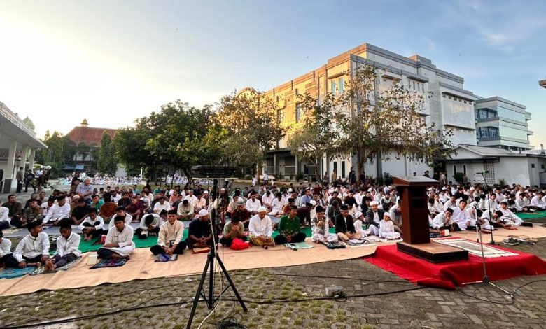 Sejumlah pria, termasuk remaja dan anak-anak, duduk rapi di atas sajadah di area parkir paving yang luas untuk sholat, dengan mimbar kayu kecil di karpet merah di depannya, di luar ruangan dekat gedung-gedung modern dan pohon di pagi hari yang cerah.