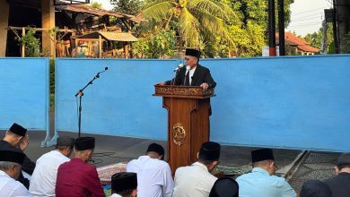 Seorang imam sekaligus khatib sedang menyampaikan khutbah Idul Adha 1447 H di atas mimbar di pelataran terbuka, disaksikan oleh jamaah pria yang duduk tertib di depannya.