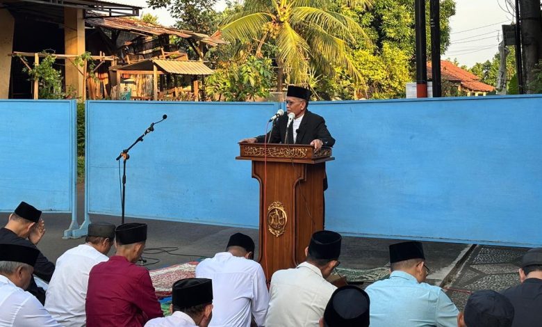 Seorang imam sekaligus khatib sedang menyampaikan khutbah Idul Adha 1447 H di atas mimbar di pelataran terbuka, disaksikan oleh jamaah pria yang duduk tertib di depannya.