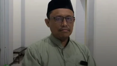 Potret foto close-up wajah Prof. Dr. Ahwan Fanani, M.Ag. mengenakan kacamata bingkai gelap, songkok hitam, dan kemeja busana Muslim hijau zaitun, melihat ke kamera dengan ekspresi tenang di latar belakang interior gedung.