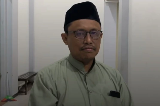 Potret foto close-up wajah Prof. Dr. Ahwan Fanani, M.Ag. mengenakan kacamata bingkai gelap, songkok hitam, dan kemeja busana Muslim hijau zaitun, melihat ke kamera dengan ekspresi tenang di latar belakang interior gedung.