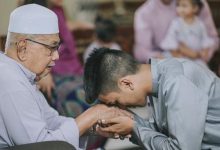 Seorang pemuda berbaju koko abu-abu sedang membungkuk mencium tangan seorang pria lansia berpeci putih dengan khidmat dalam suasana kekeluargaan.