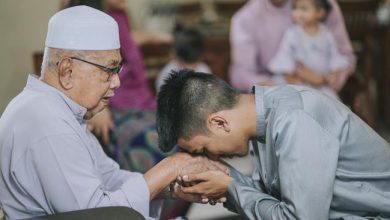 Seorang pemuda berbaju koko abu-abu sedang membungkuk mencium tangan seorang pria lansia berpeci putih dengan khidmat dalam suasana kekeluargaan.